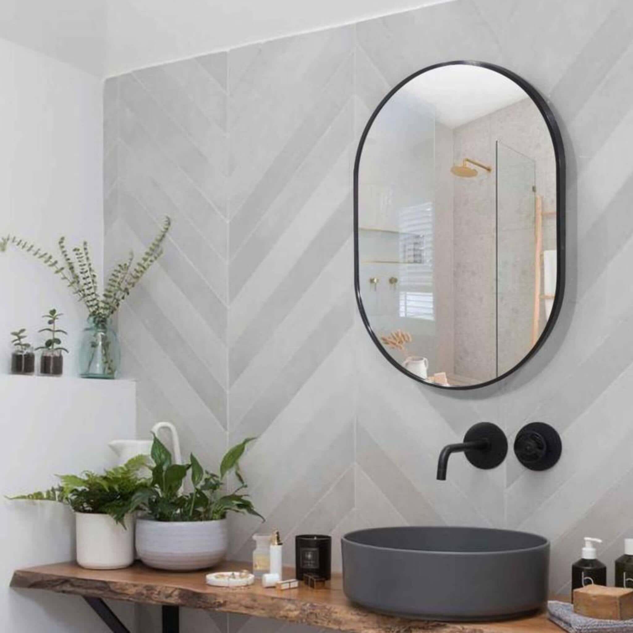 Modern small oval black aluminium mirror above a bathroom sink with gray herringbone wall and greenery.