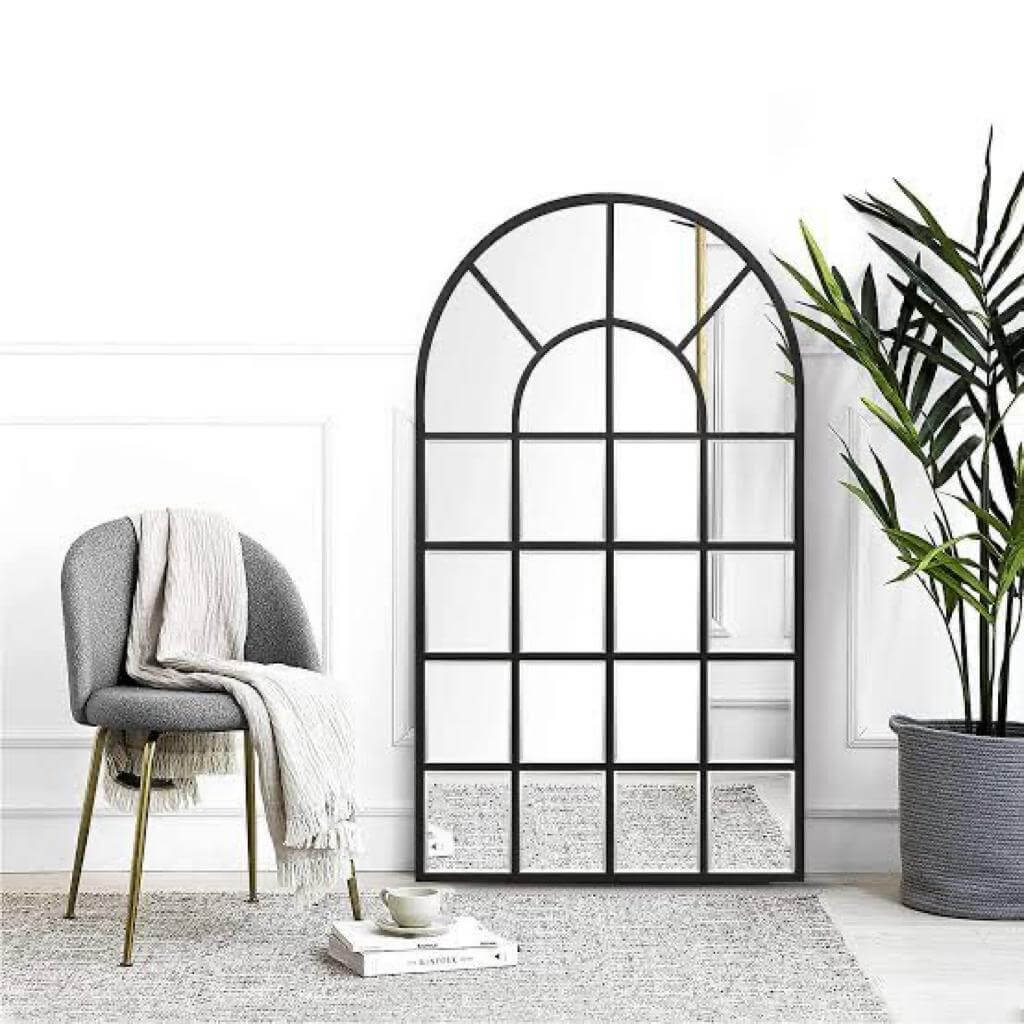 Large arched mirror with window panel design next to a modern chair and plant, ideal for home decor.