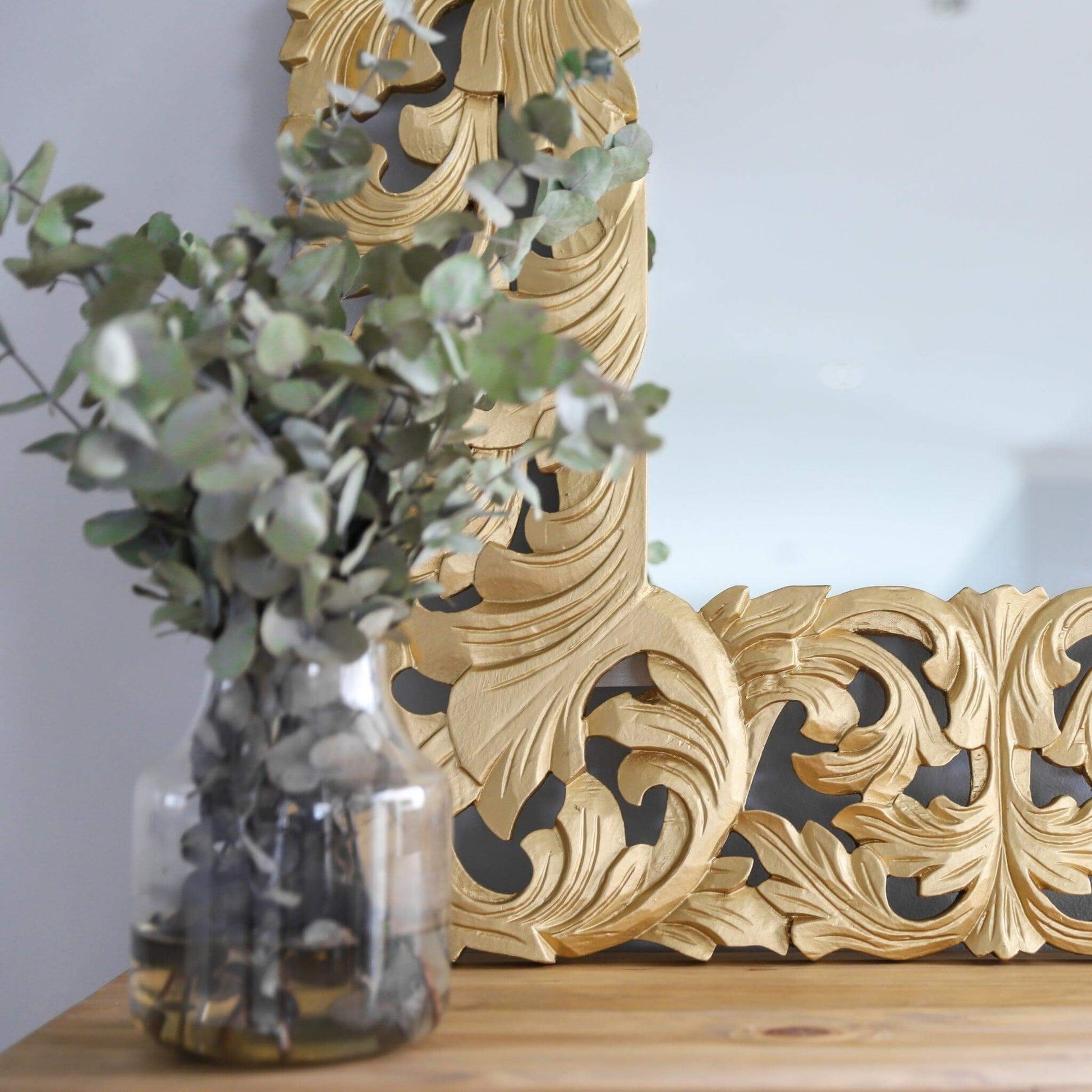 Decorative gold ornate frame of the Envy Mirror with a vase of eucalyptus on a wooden surface.