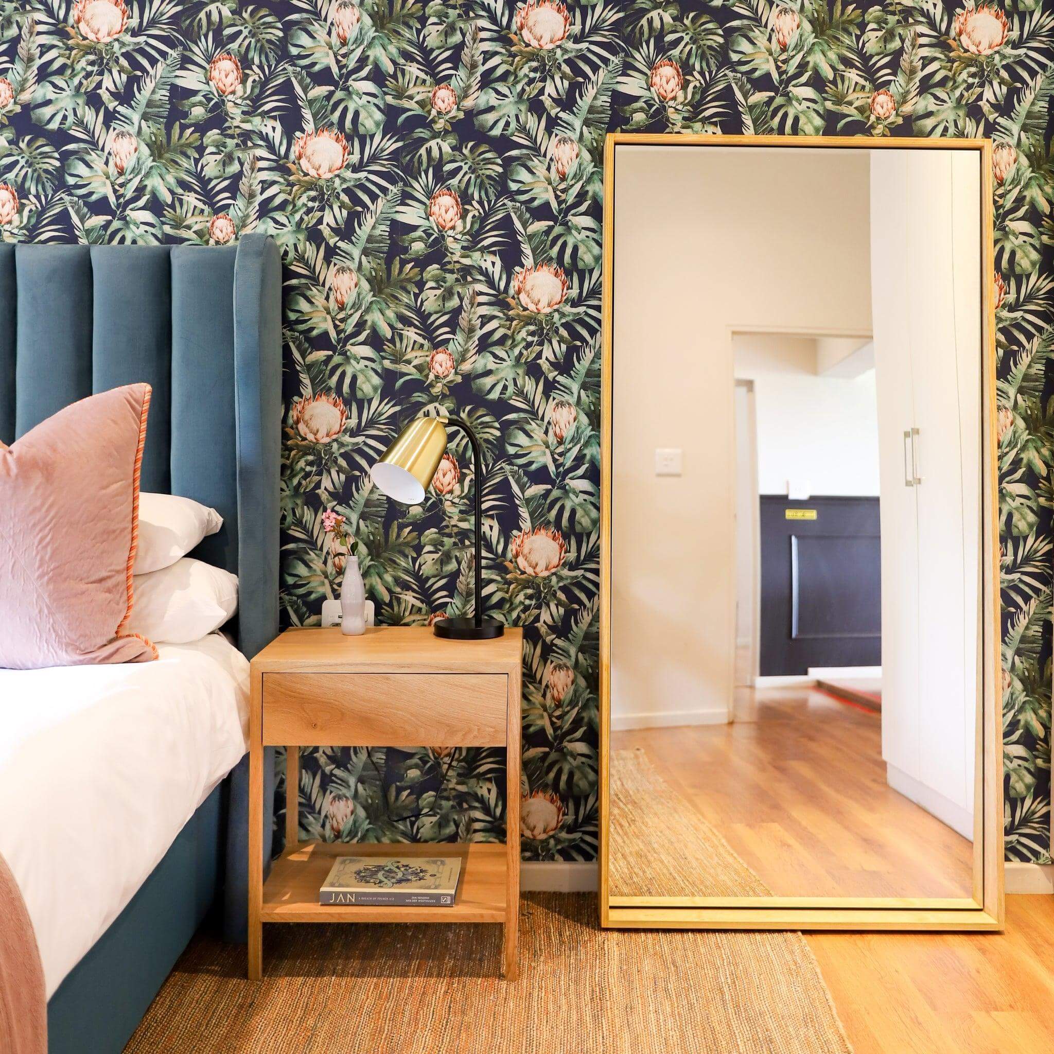 Jupiter Mirror leaning against a floral wall in a modern bedroom, showcasing its elegant wood-styled frame.